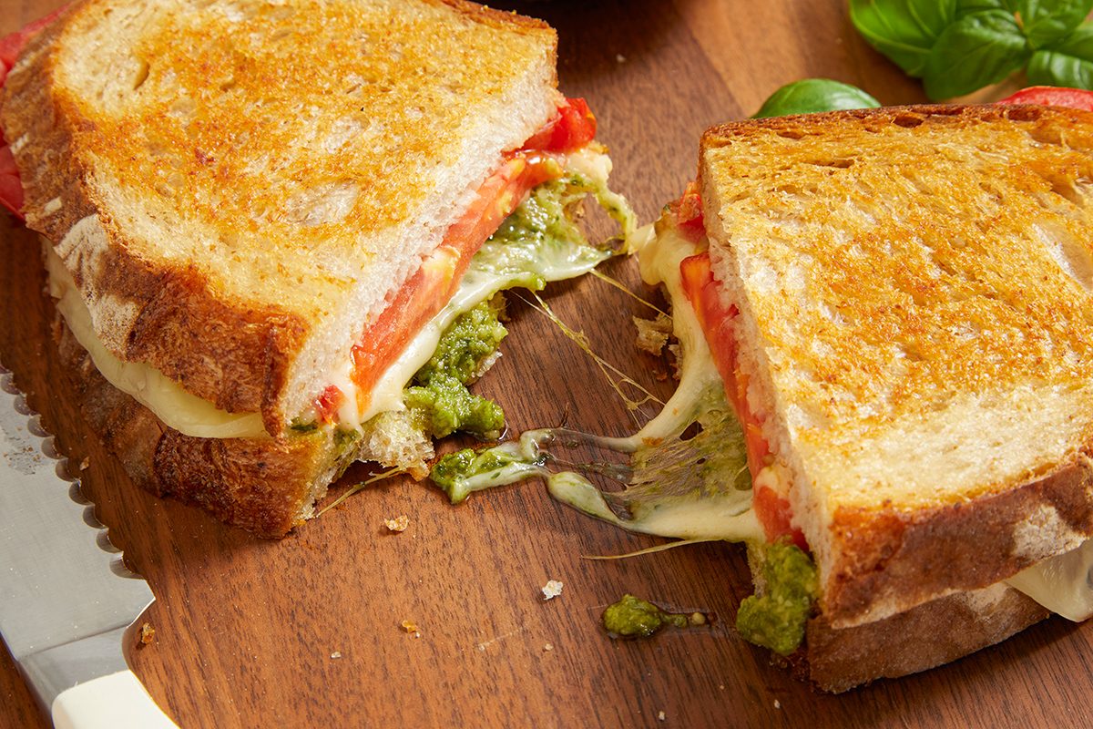 A grilled cheese sandwich cut in half on a wooden board, with melted cheese, tomato slices, and green pesto spilling out. A serrated knife and fresh basil leaves are nearby.
