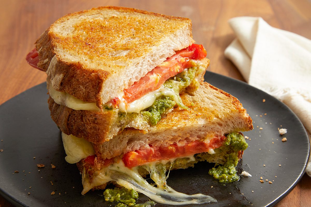 A grilled cheese sandwich with melted cheese, tomatoes, and green pesto, stacked on a black plate with a white napkin in the background.
