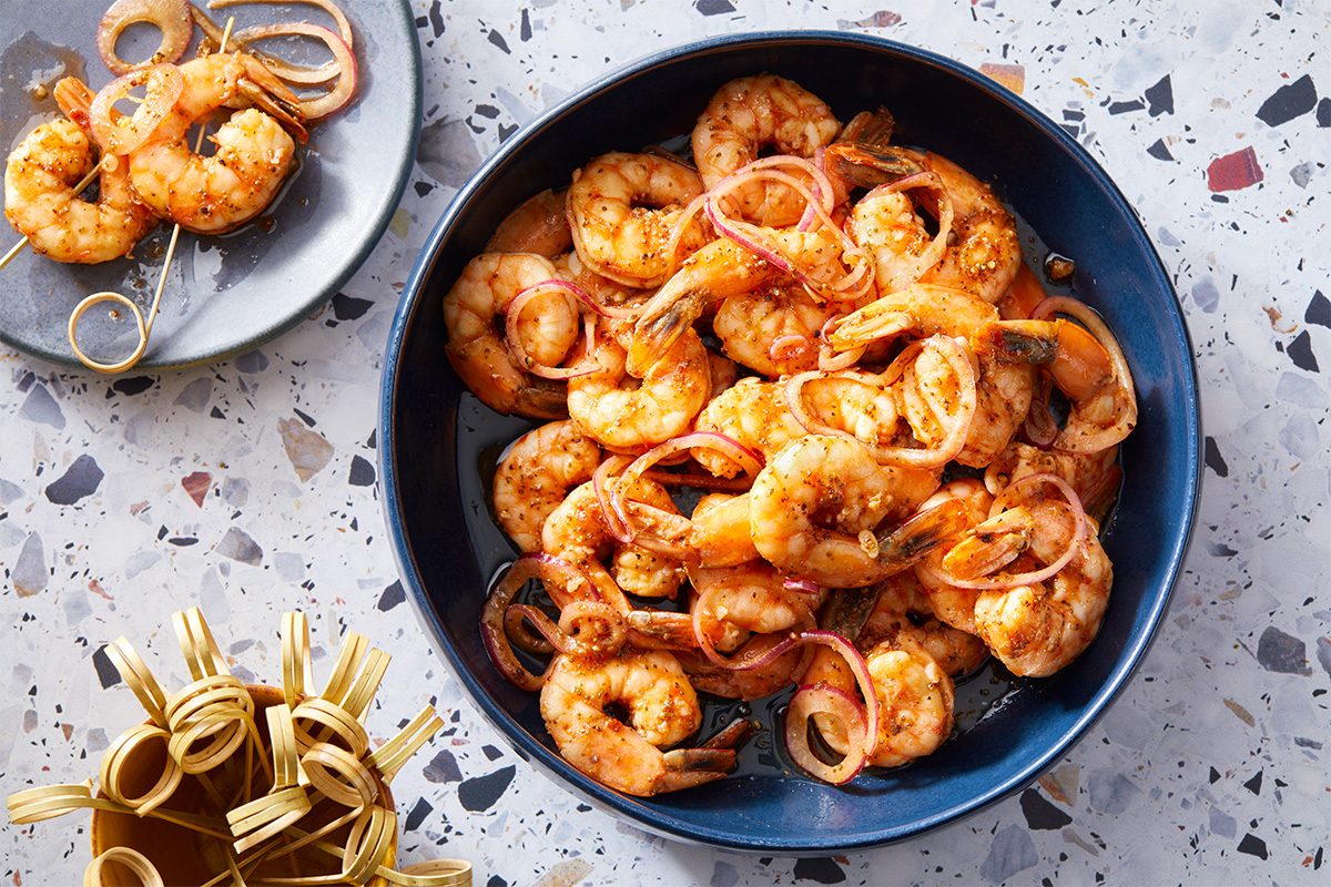 A horizontal overhead view shows a bowl of shrimp alongside small dishes and garnishes arranged on a tabletop.