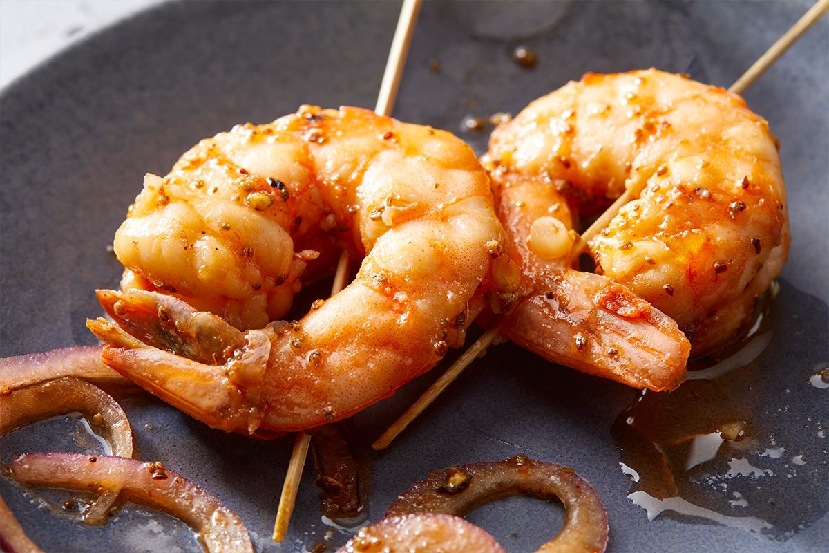 A horizontal close-up shows two shrimp skewered on sticks with visible seasoning and glaze.