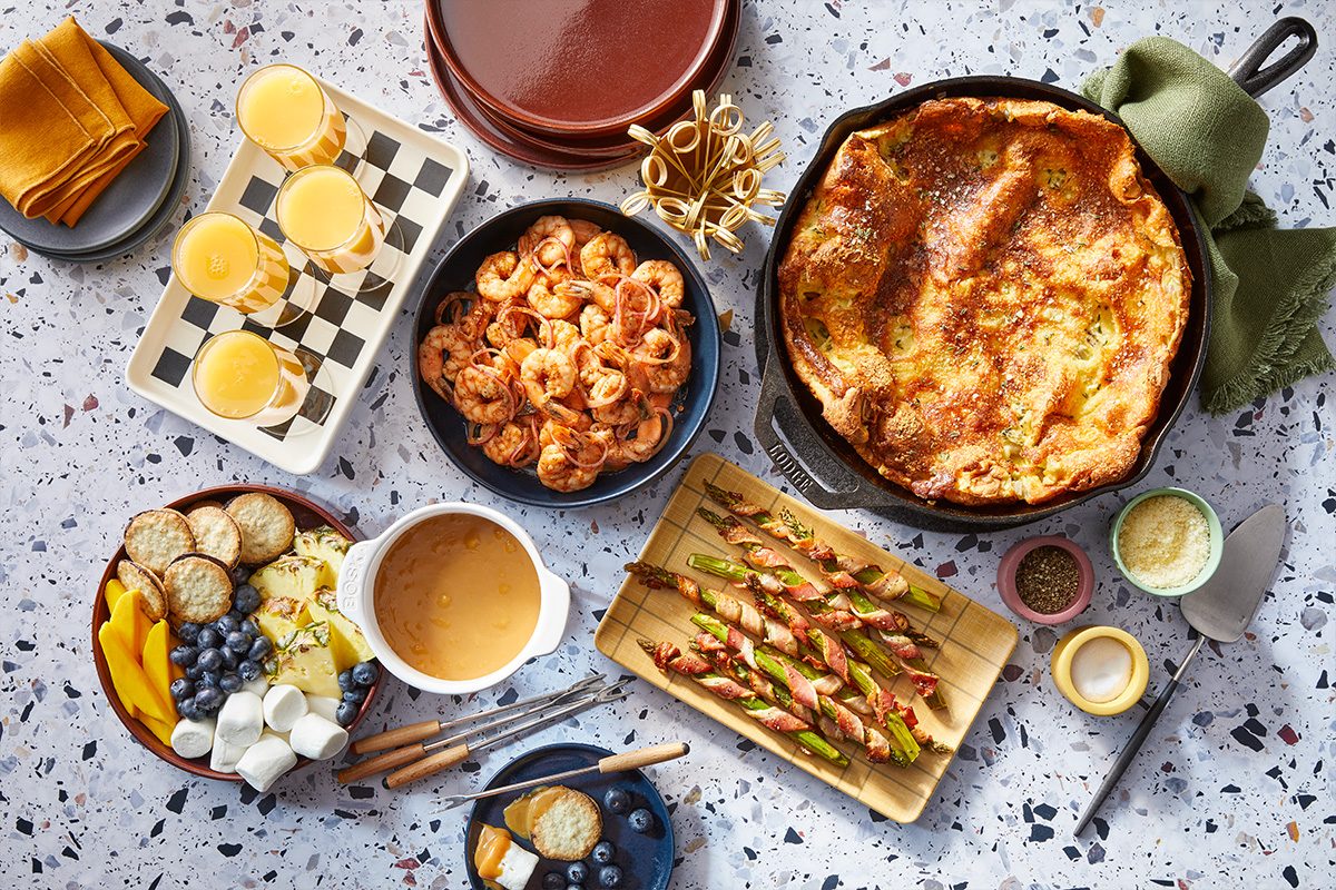 A horizontal overhead image shows multiple brunch dishes including shrimp, Dutch baby, asparagus, mimosas, and dulce de leche arranged together.