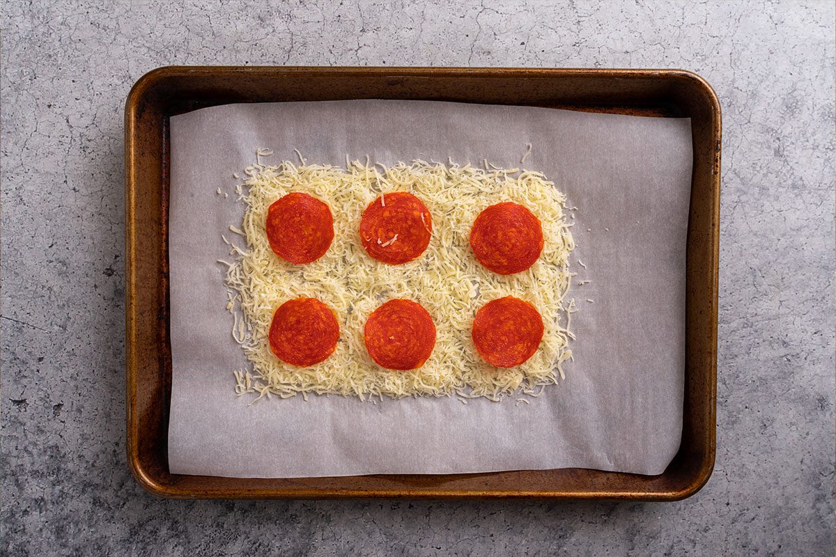 Unbaked pepperoni pizza base made with shredded cheese spread on parchment paper and topped with six slices of pepperoni on a baking tray.