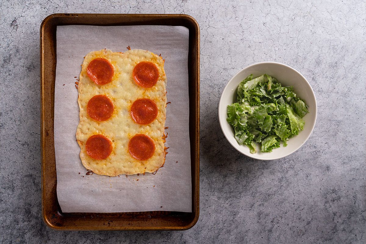 Baked cheese and pepperoni crust on a parchment-lined tray with a small bowl of Caesar salad on the side.