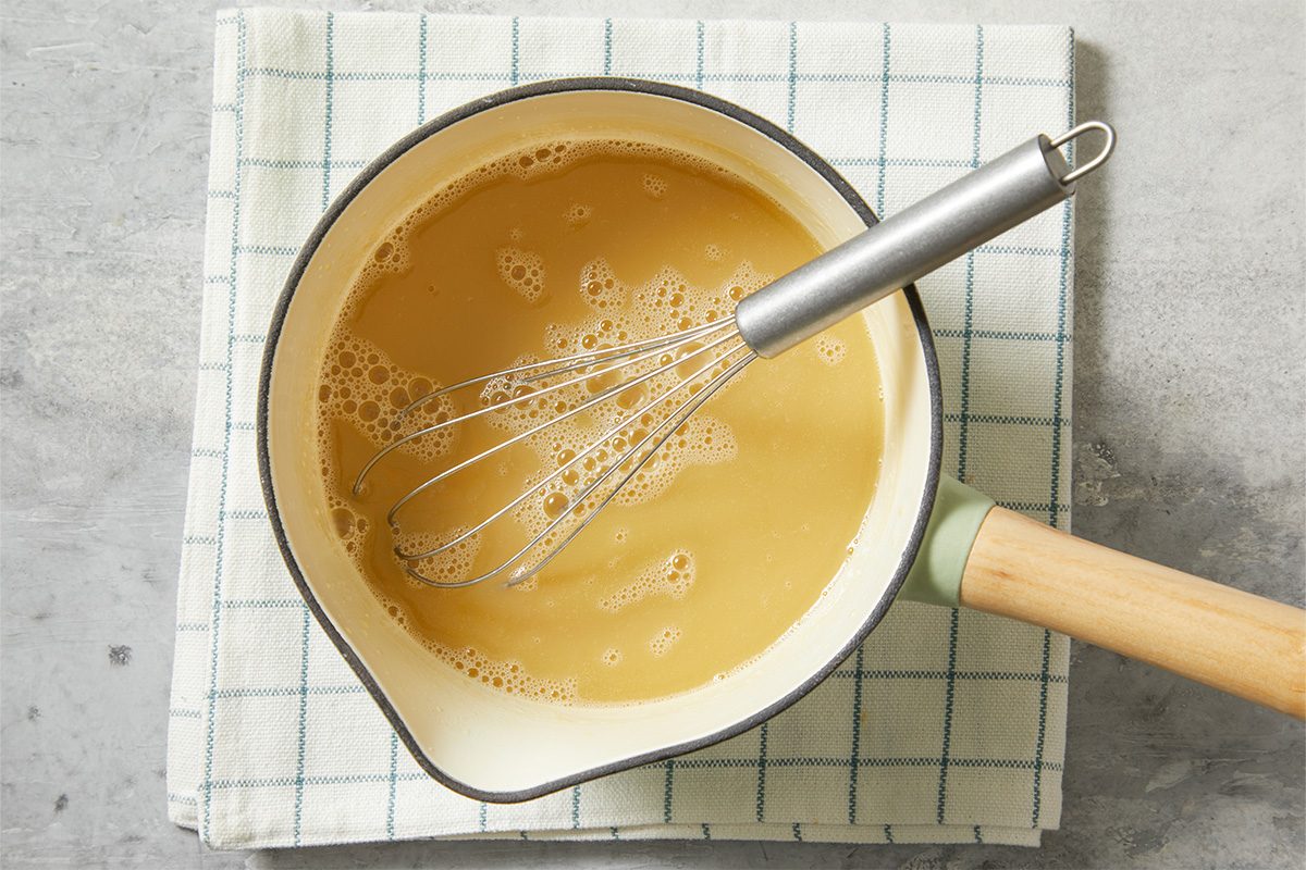 A horizontal overhead image shows a saucepan filled with a maple cream mixture being whisked on a stovetop surface.