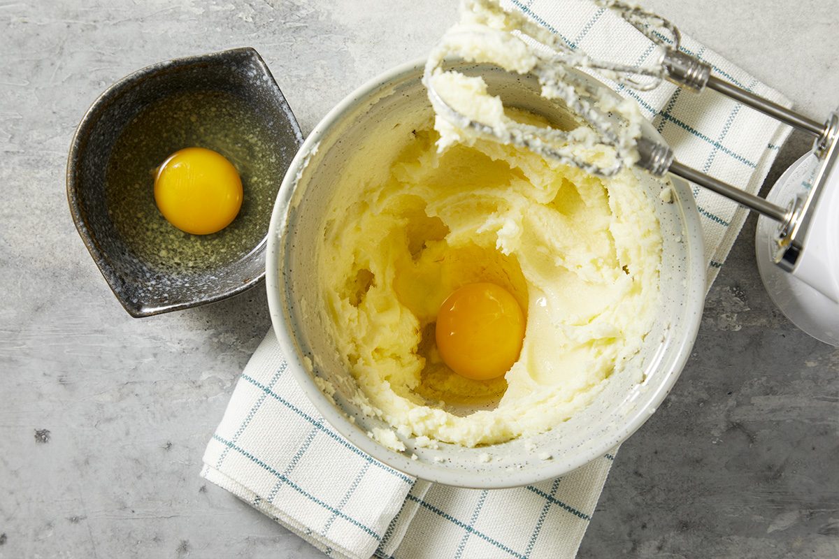 A horizontal overhead image shows softened butter being mixed with eggs in a stand mixer bowl with additional ingredients nearby.