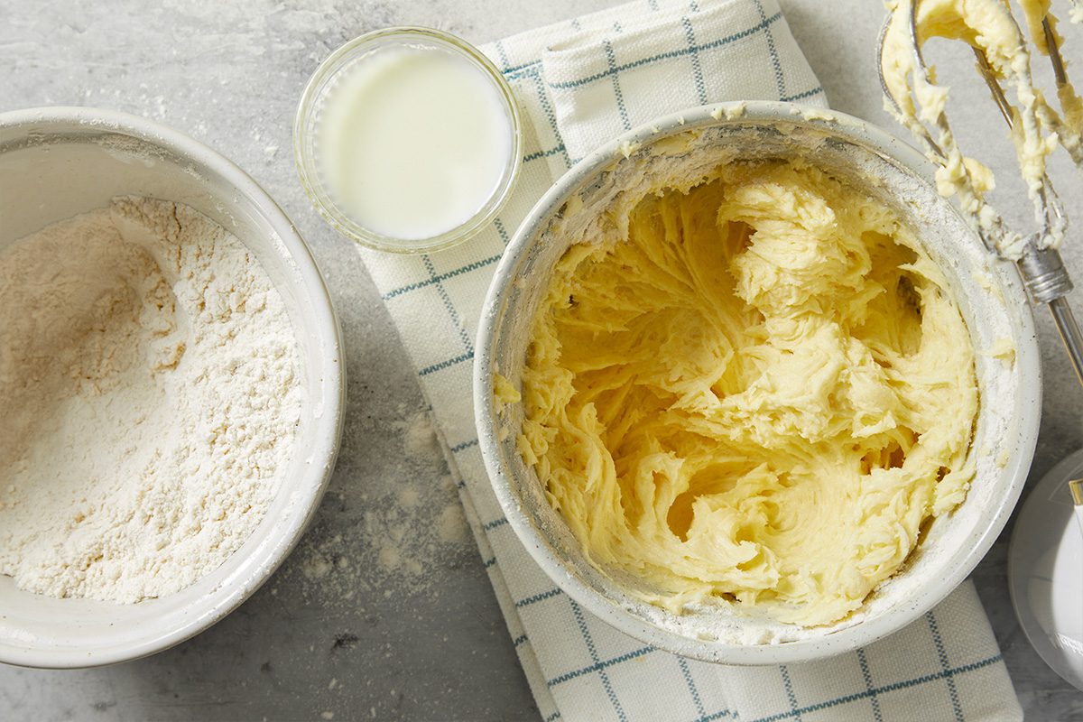 A horizontal overhead image shows flour and milk being added to a creamed butter mixture in a mixing bowl.