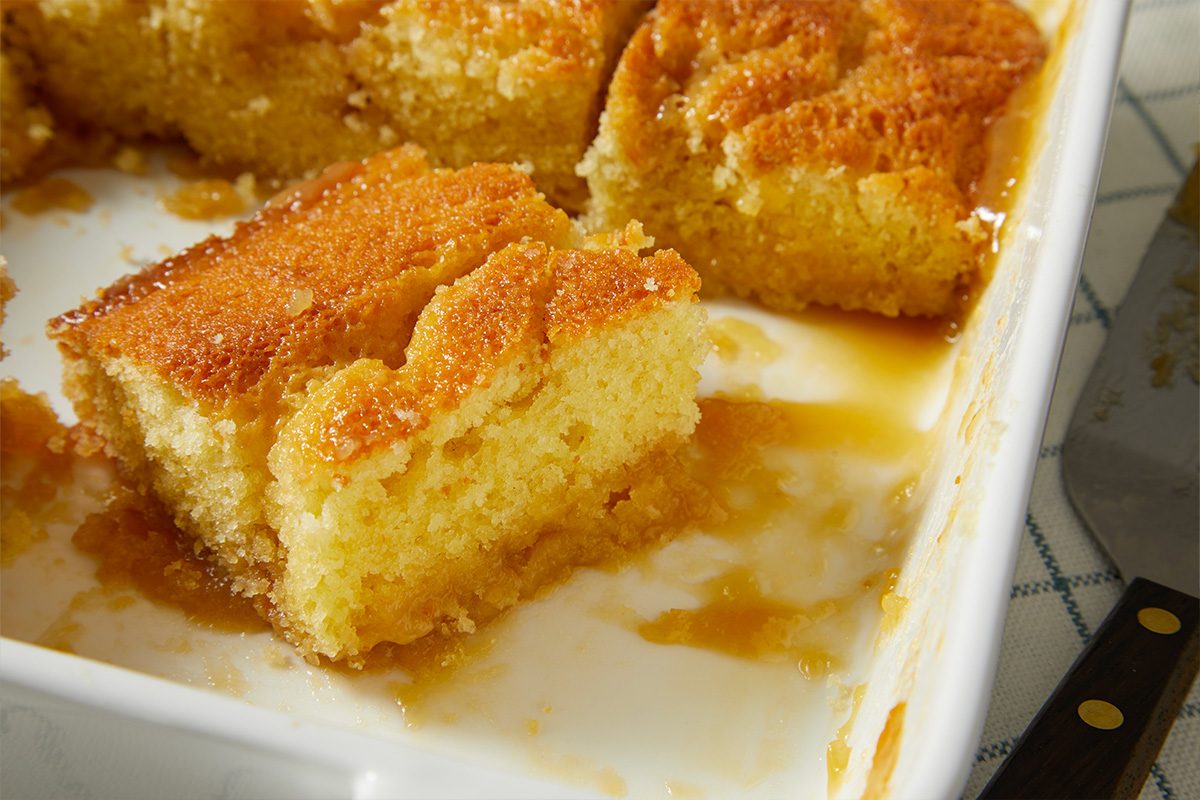 A horizontal close-up image shows a single square of pudding with a soft crumb and sauce soaking into the base.