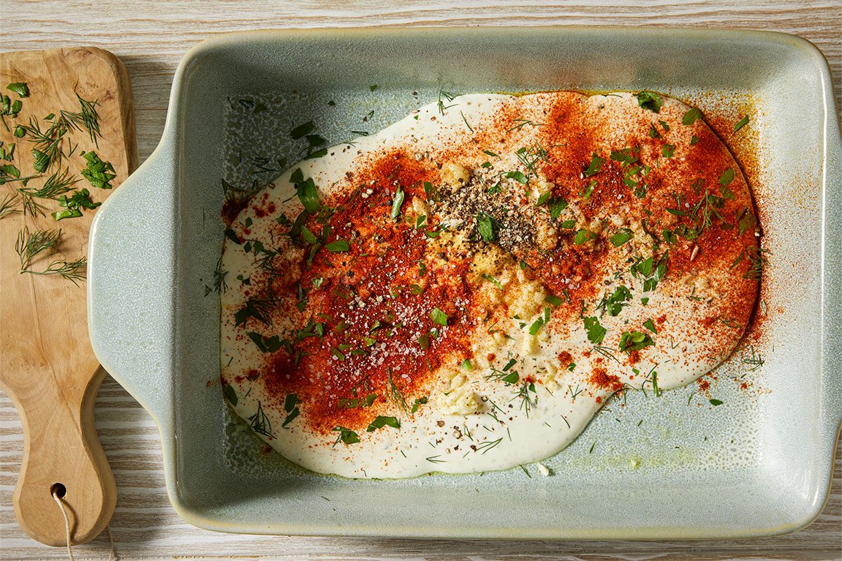 A horizontal overhead image shows a creamy ranch-based mixture in a baking dish topped with paprika, herbs, and seasonings, with a wooden board and fresh dill nearby.