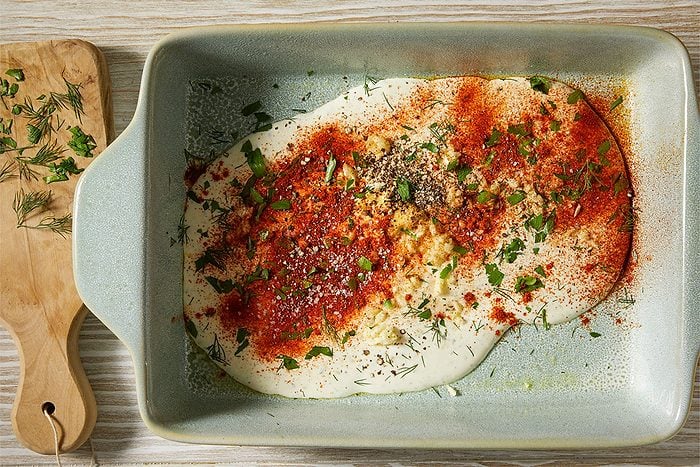 A horizontal overhead image shows a creamy ranch-based mixture in a baking dish topped with paprika, herbs, and seasonings, with a wooden board and fresh dill nearby.