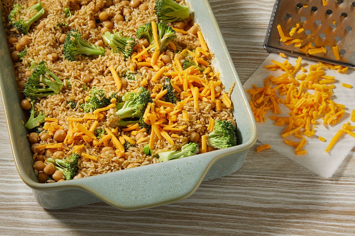A horizontal overhead image shows a baking dish filled with brown rice, chickpeas, broccoli, and shredded cheese, with additional cheese scattered beside a grater.