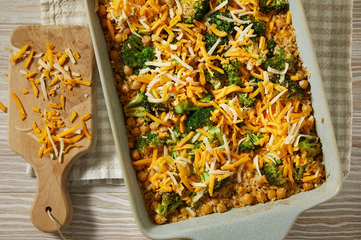 A horizontal overhead image shows a casserole layered with chickpeas, broccoli, and shredded cheese, with a wooden spoon and extra cheese nearby.