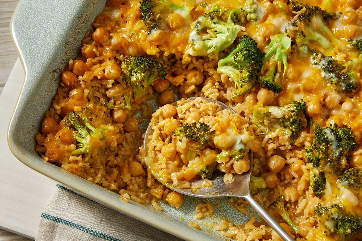 A horizontal close-up shows a spoon scooping a portion of casserole with chickpeas, rice, broccoli, and melted cheese from the baking dish.