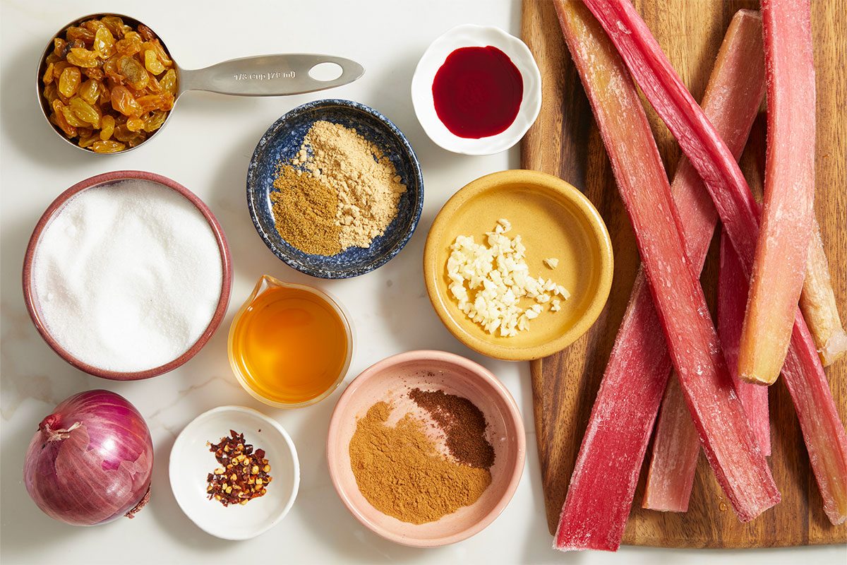 Overhead ingredient shot of chopped rhubarb, spices, sugar, vinegar, onion, and raisins arranged for chutney preparation.
