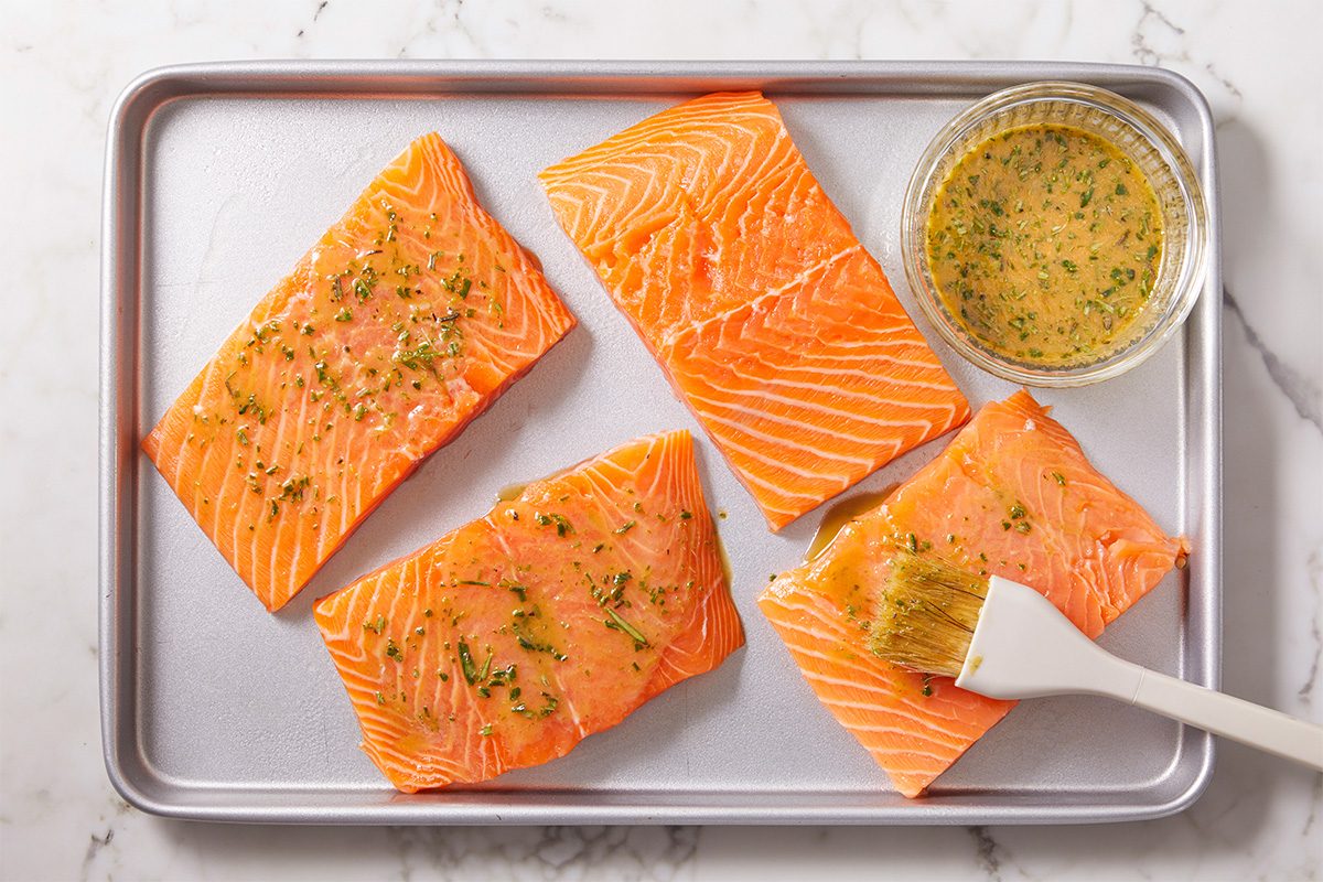 Overhead horizontal image of salmon fillets brushed with herb marinade on a sheet pan with sauce nearby.