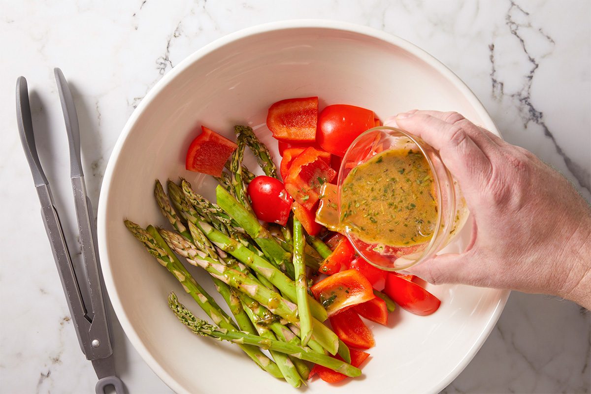 Overhead horizontal image of asparagus and red pepper being tossed with marinade in a mixing bowl.