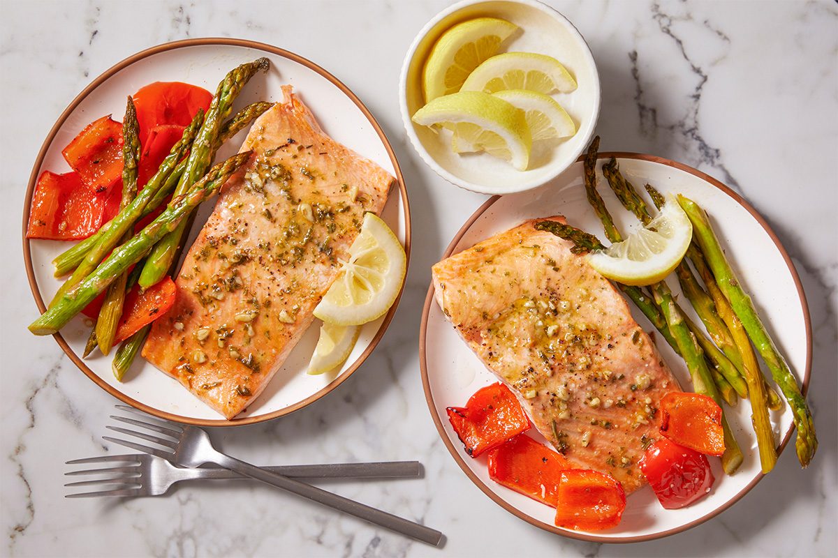 Overhead horizontal image of plated salmon fillets with asparagus, peppers, and lemon wedges.