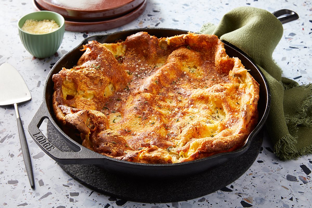 A golden-brown Dutch baby pancake rises high in a cast iron skillet, sitting on a speckled countertop with a green cloth and a small bowl of grated cheese nearby.