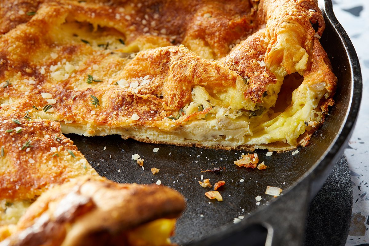 A golden-brown Dutch baby pancake with a puffy, crispy edge sits in a black skillet. A slice has been cut out, revealing a fluffy, airy interior with a light dusting of powdered sugar on top.