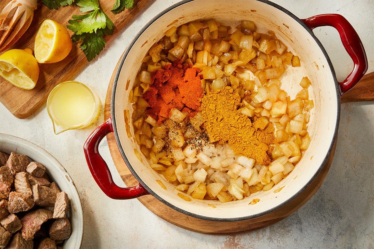 Overhead horizontal image of onions and spices including curry powder and garam masala being cooked with butter in a Dutch oven with cubed beef nearby.
