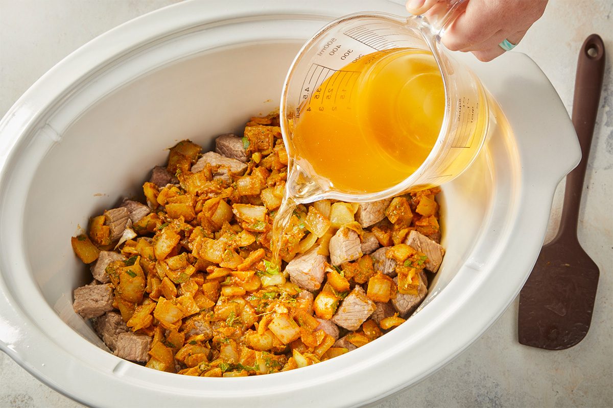 Overhead horizontal image of beef, vegetables, and spices in a slow cooker as broth is poured over the mixture.