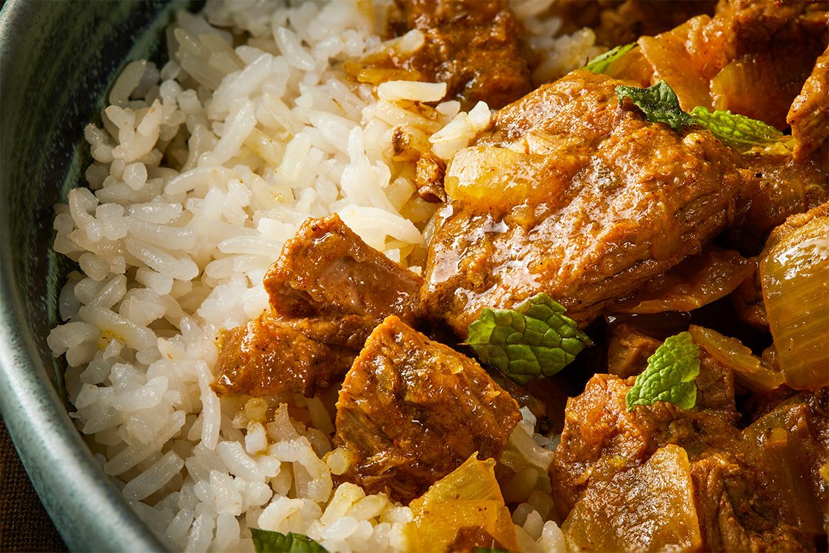 Overhead horizontal close-up of curried beef stew over rice showing texture of sauce, onions, and tender beef pieces.