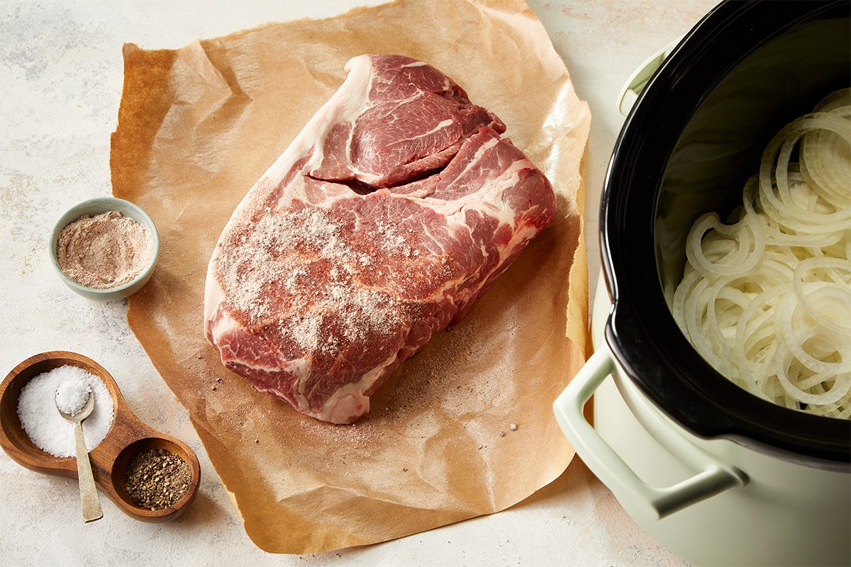 Overhead horizontal image of raw pork shoulder placed on parchment with sliced onions and seasonings beside a slow cooker.. COMBO SHOT; onions placed in slow cooker, pork next to slow cooker on butcher paper or cutting board, being sprinkled withs seasonings.