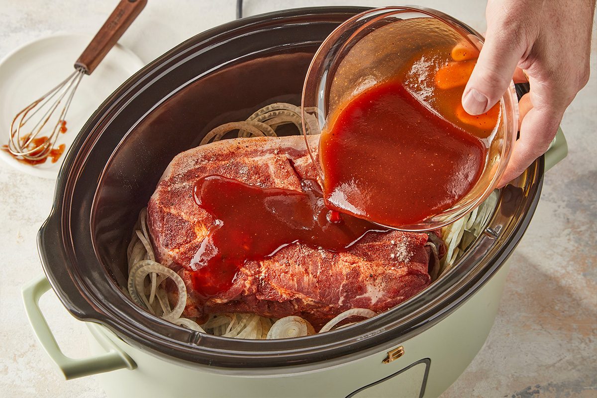 Overhead horizontal image of barbecue sauce mixture being poured over pork and onions inside a slow cooker.. Pouring sauce ingredients over pork.