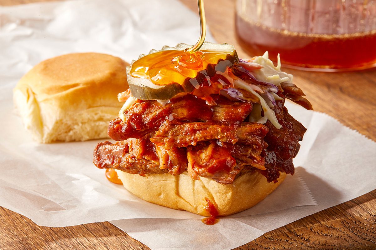 Eye-level horizontal image of a pulled pork slider topped with barbecue sauce and pickles on a soft bun. Assembly shot; drizzling hot honey over pork on slider bun, coleslaw nearby.