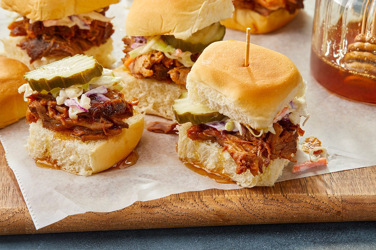 Eye-level horizontal image of pulled pork sliders served on a wooden board with pickles and coleslaw.