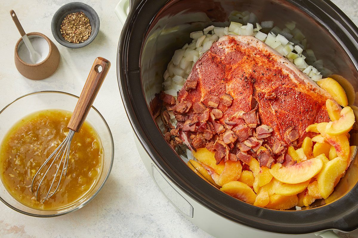 Overhead horizontal image of pork, peaches, bacon, and sauce ingredients assembled inside a slow cooker for peach and green chile pork. Seasoned pork in slow cooker. Top with onions, peaches, and bacon. Small bowl with enchilada sauce, green chiles, bbq sauce, and garlic.