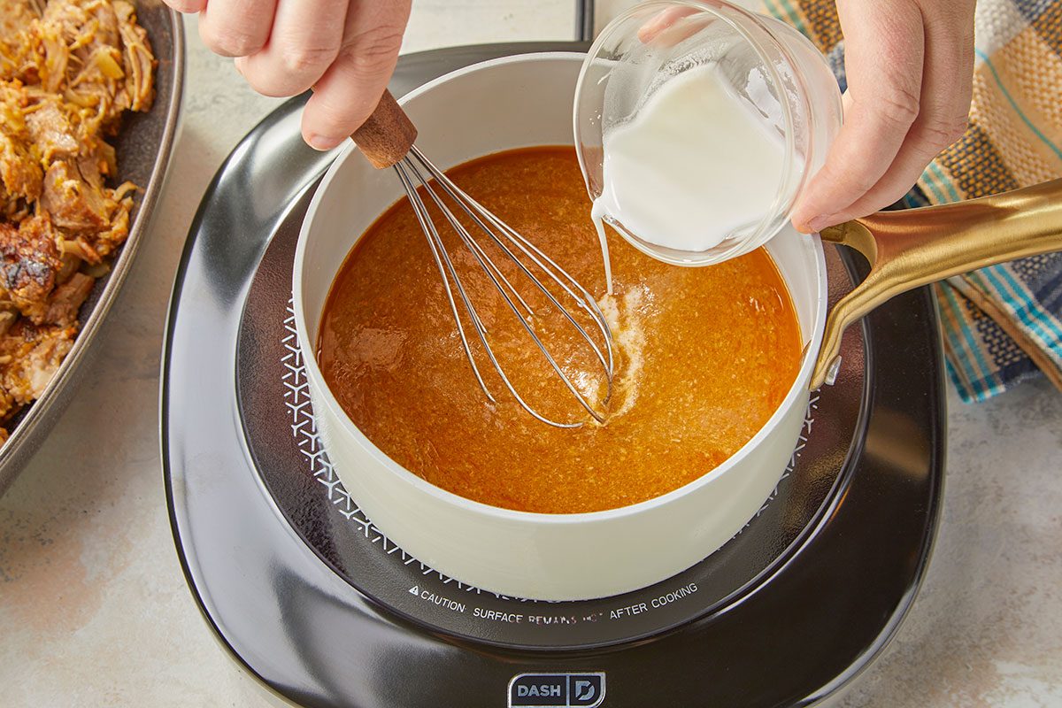 Overhead horizontal image of sauce being whisked together for slow cooker peach and green chile pork.. Small saucepan with cooking liquid. Small glass measuring cup with cornstarch and water. Whisk into cooking liquid.