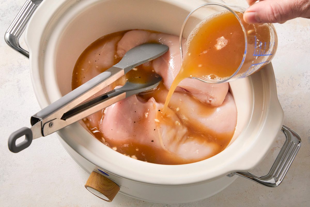 broth, pineapple juice, garlic, salt and pepper mixture being poured over chicken in a slow cooker