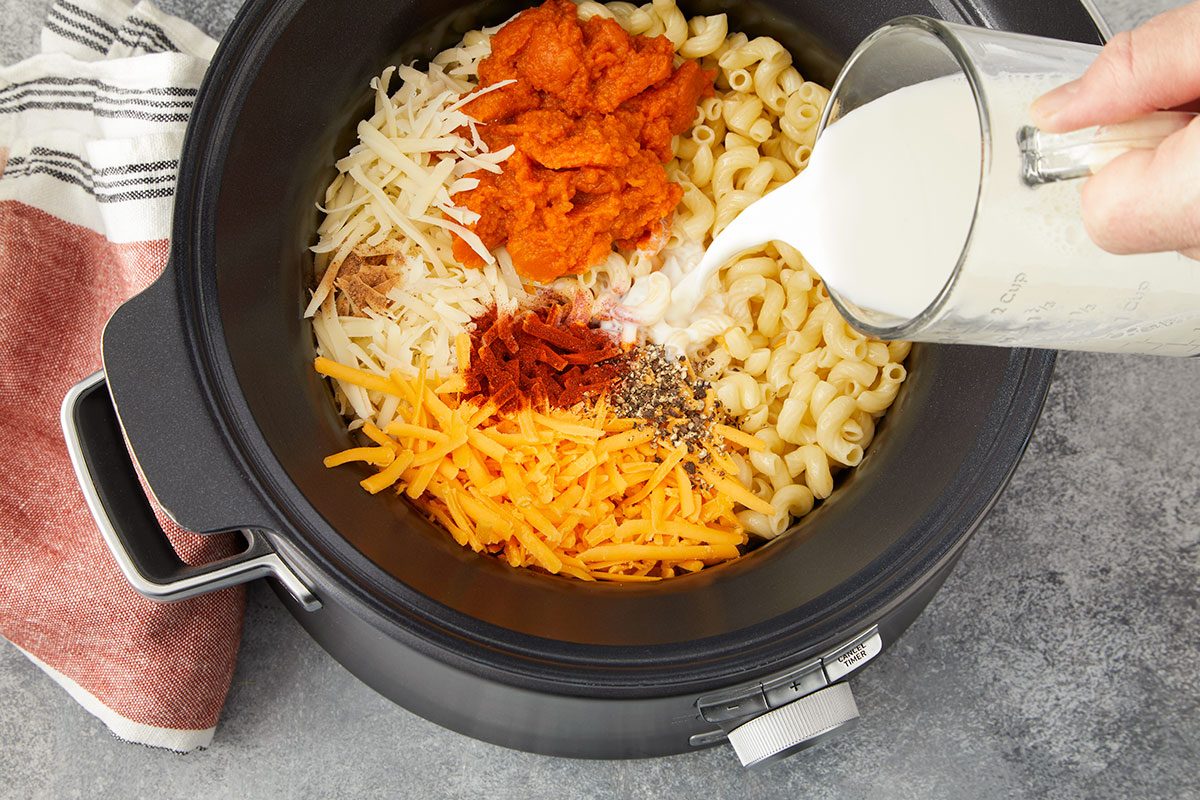 Overhead horizontal image of elbow macaroni in a slow cooker with pumpkin puree, shredded cheeses, spices, and milk being poured in.. Step 1: Pouring milk over pasta, cheeses and other ingredients in the slow cooker