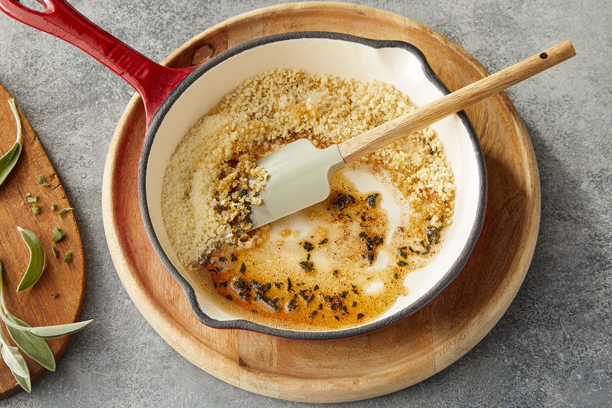 Overhead horizontal image of browned butter with chopped sage and toasted breadcrumbs being prepared in a skillet.. Step 3: Stirring panko and Parmesan into skillet with butter and cooked sage leaves