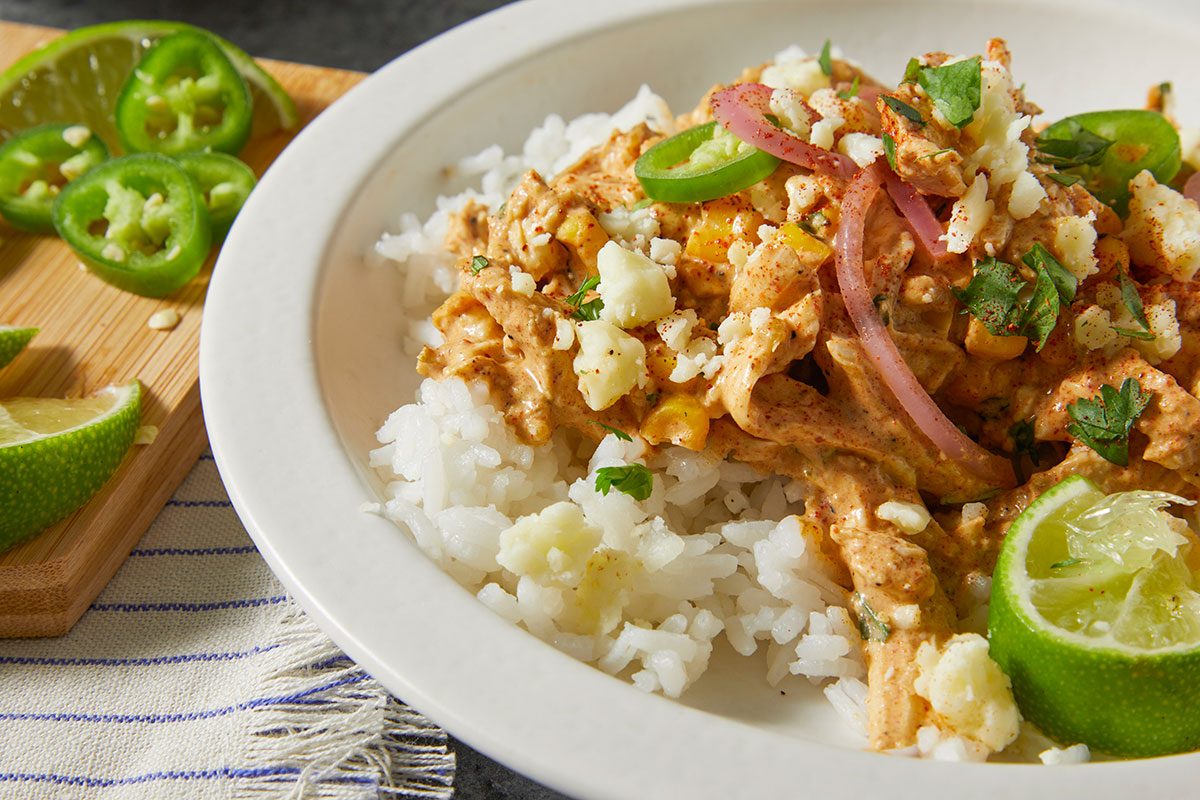 Overhead horizontal push and pulls image of creamy street corn chicken served over rice with fresh garnishes and lime.