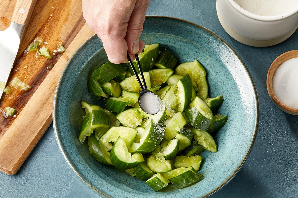 A hand sprinkles salt from a spoon onto chopped cucumbers in a blue bowl. A wooden cutting board with cucumber pieces and a small cup of salt are nearby on a blue surface.