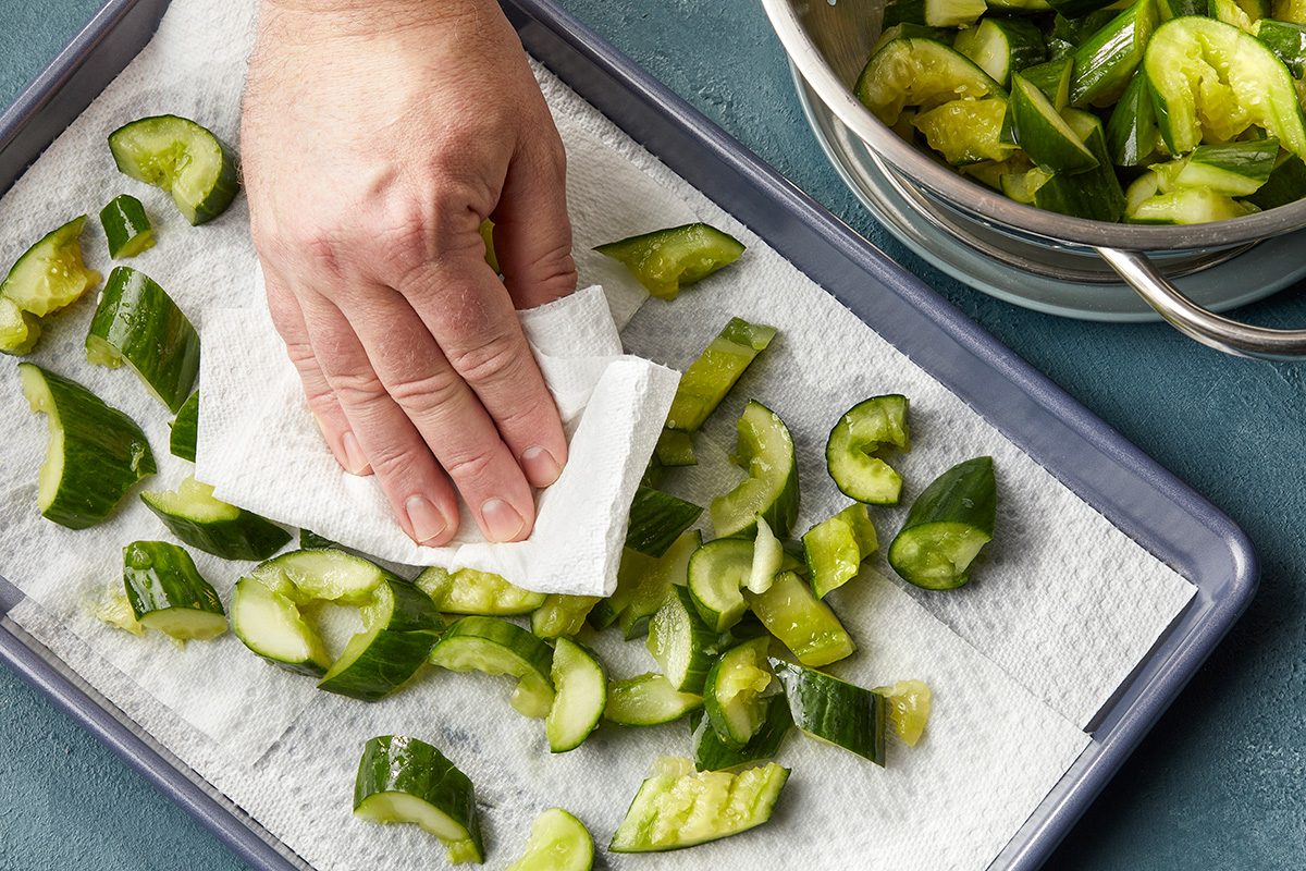 A hand presses a paper towel onto chopped cucumbers spread on a paper towel-lined baking sheet, with a bowl of more cucumbers nearby.