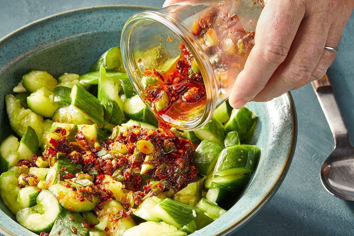 A hand pours a spicy red chili oil mixture over chopped cucumbers in a blue bowl, with a spoon resting beside the bowl.