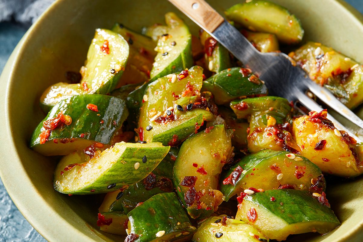 A bowl of sliced cucumbers mixed with red chili flakes and sesame seeds, served with a fork. The cucumbers appear to be lightly seasoned and tossed in a flavorful dressing.