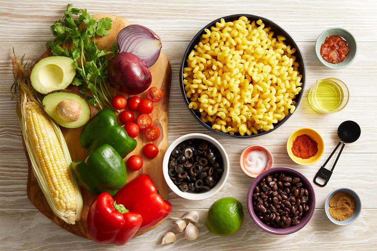 Overhead shot of ingredients for Southwestern Spiral Pasta, cooked pasta, corn, bell peppers, black beans, tomatoes, red onion, and lime arranged on a white surface.