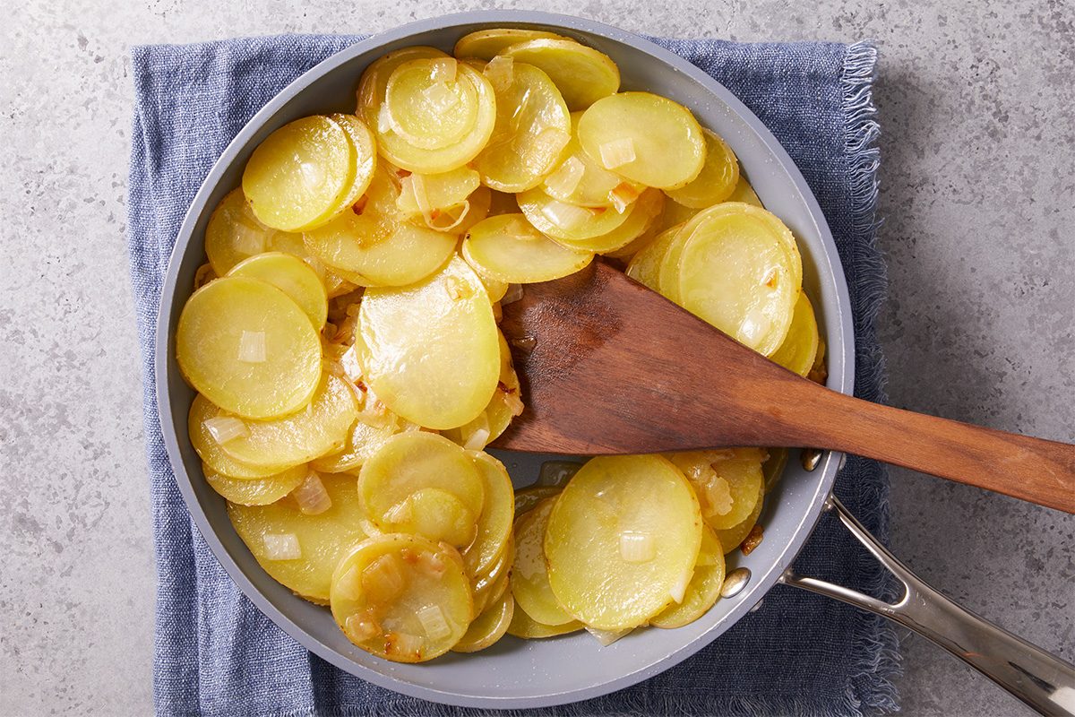 Overhead step-by-step image of sliced Yukon Gold potatoes sautéing in olive oil in a skillet with a wooden spoon, beginning the cooking process for an Easy Spanish Omelet on a neutral countertop.