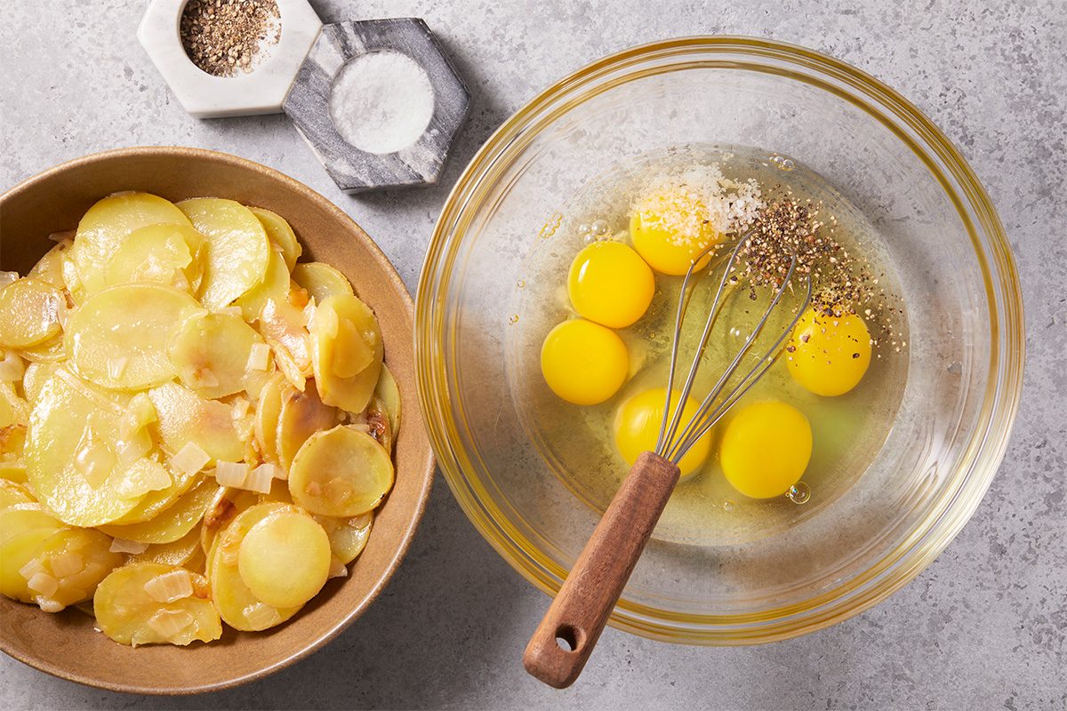 Overhead step-by-step preparation image showing eggs being whisked in a glass bowl with salt and pepper beside sautéed potatoes and chopped onion for an Easy Spanish Omelet.