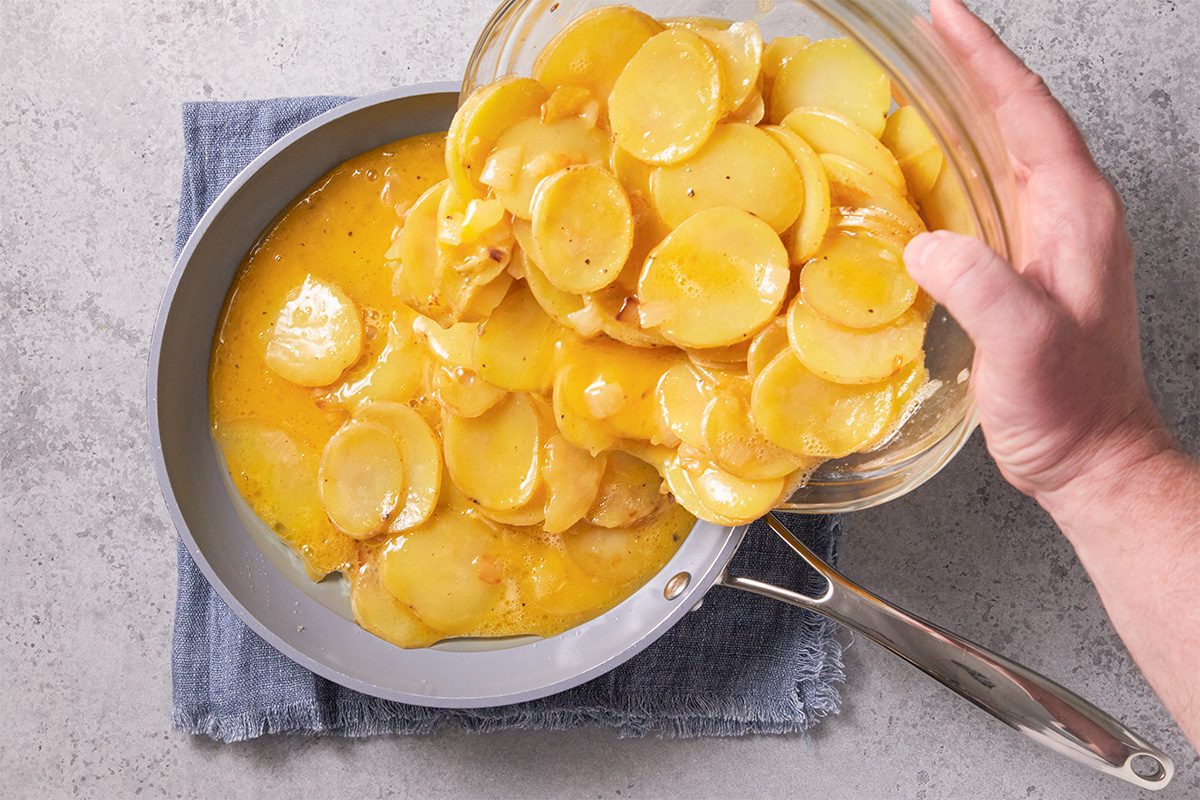 Overhead step-by-step cooking image of potato and egg mixture being poured into a skillet to form an Easy Spanish Omelet, showing sliced potatoes coated in egg mixture.