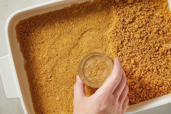 Graham cracker crumbs mixed with butter and sugar are pressed firmly into a baking dish to form the crust.