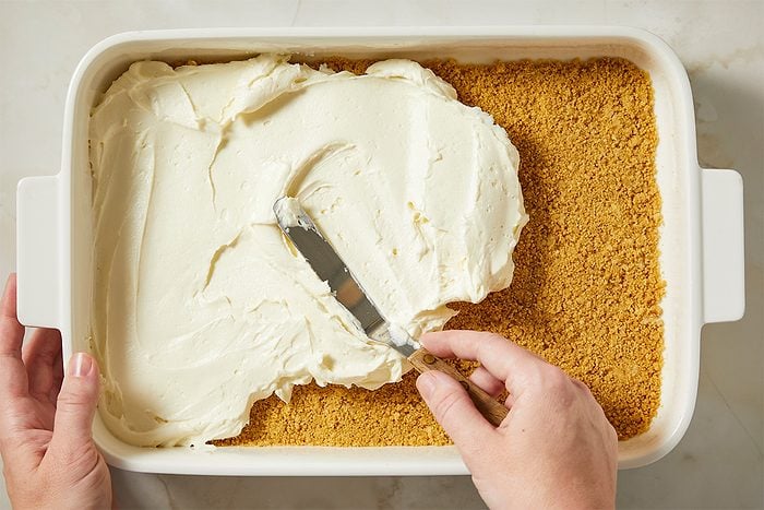 The cream cheese filling is spread evenly over the graham cracker crust using a spatula.
