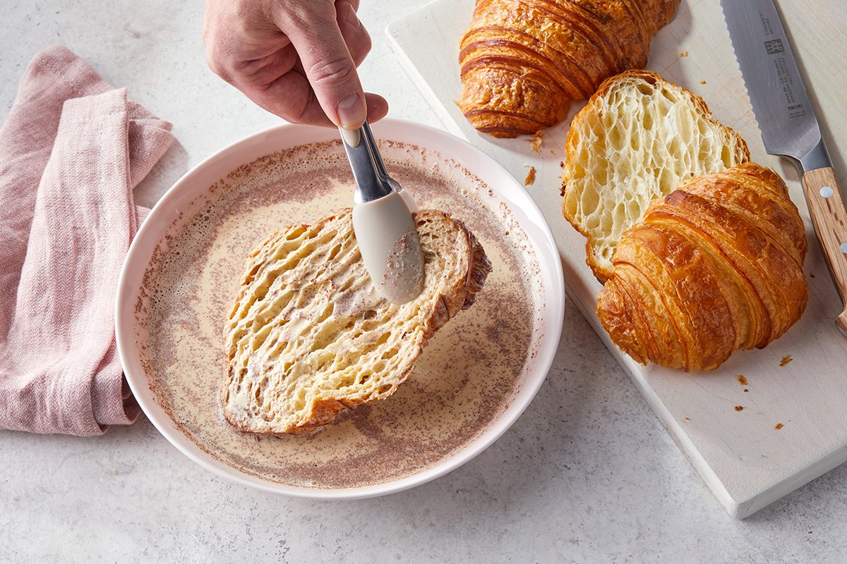 A hand dips a sliced croissant into a bowl of beaten eggs or batter. Nearby, more croissant slices and a knife rest on a white cutting board. A pink napkin is placed to the side.