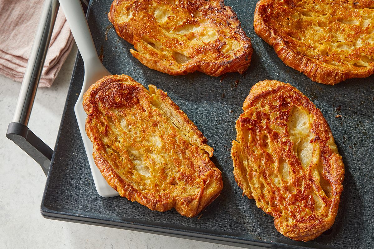 Golden brown slices of French toast are cooking on a stovetop griddle, with a spatula lifting one of the pieces. The toast appears crispy and well-cooked with a slightly fluffy texture.