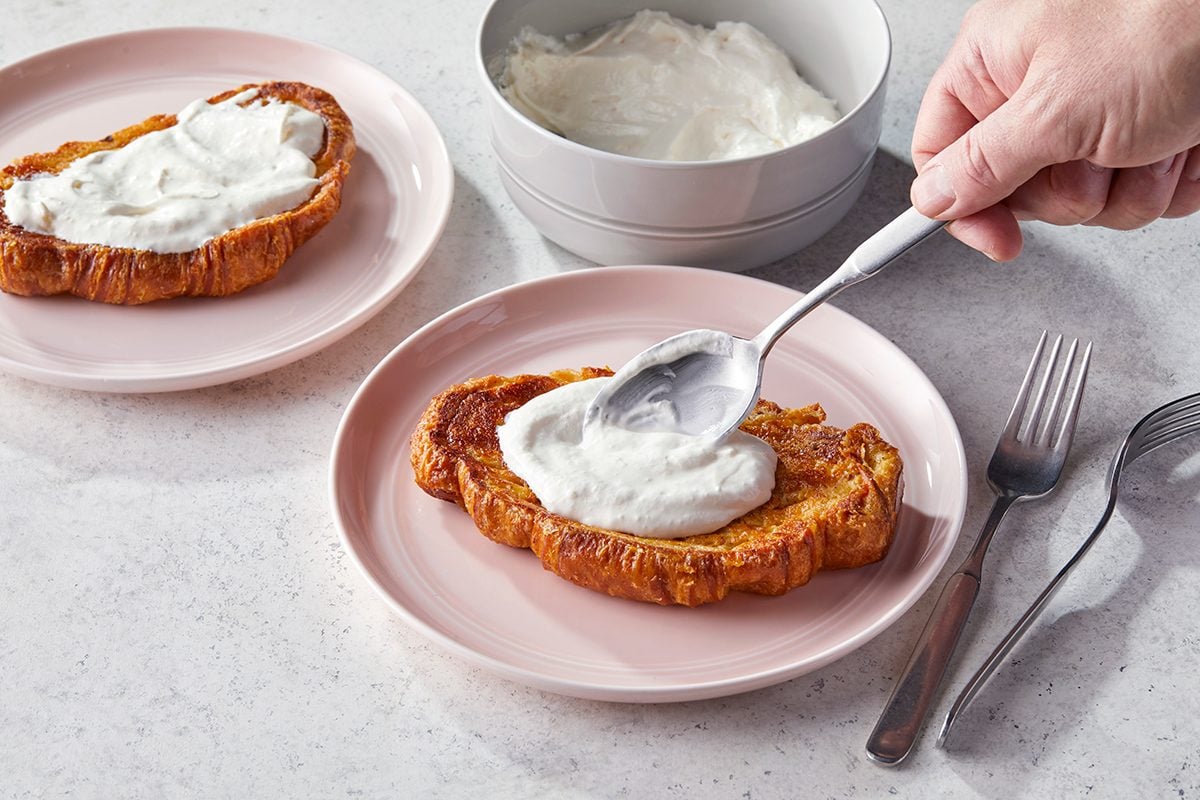 A hand spreads a dollop of whipped cream or yogurt onto a toasted croissant slice on a pink plate. Another plate with a prepared croissant and a bowl of cream are nearby, with two forks beside the plate.