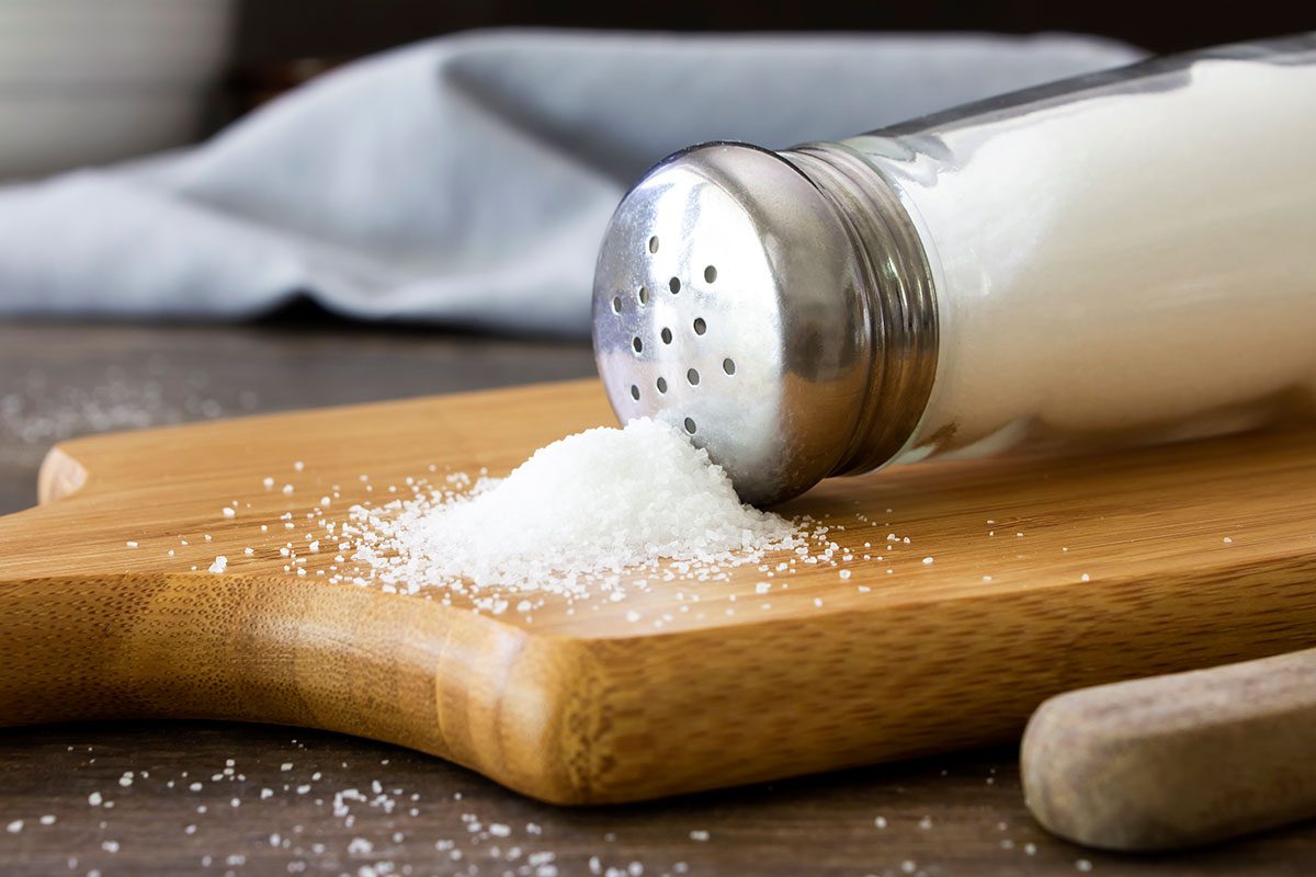 A close up shot of salt spilling out of a salt shaker on to a wooden chopping board.