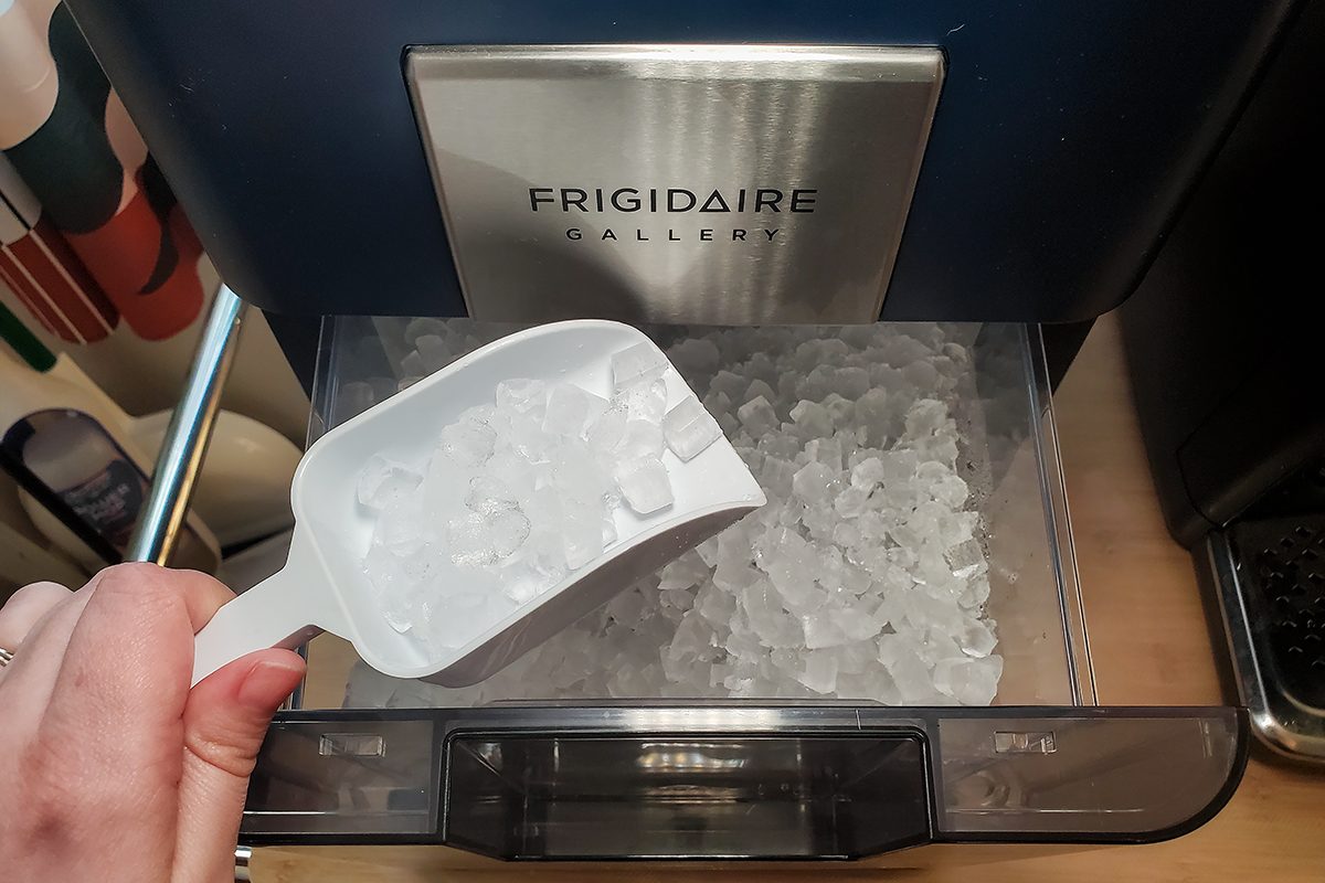A hand holding a white plastic scoop filled with ice cubes above a bin of ice inside a Frigidaire Gallery ice maker.
